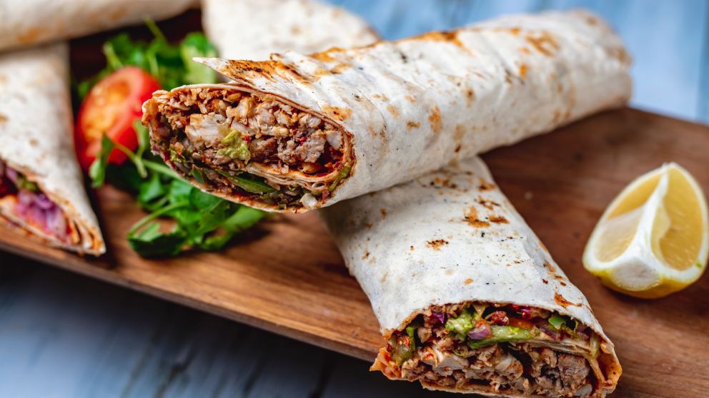 side view chicken doner with greens cucumber tomato sauce cucumber red onion in pita and slice of lemon on a board