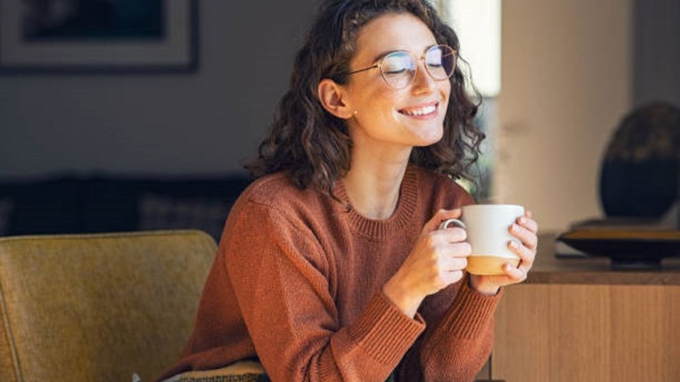 Happy young woman drinking a cup of tea in an autumn morning. Dreaming girl sitting in living room with cup of hot coffee enjoying under blanket with closed eyes. Pretty woman wearing sweater at home and enjoy a ray of sunshine on a winter afternoon.