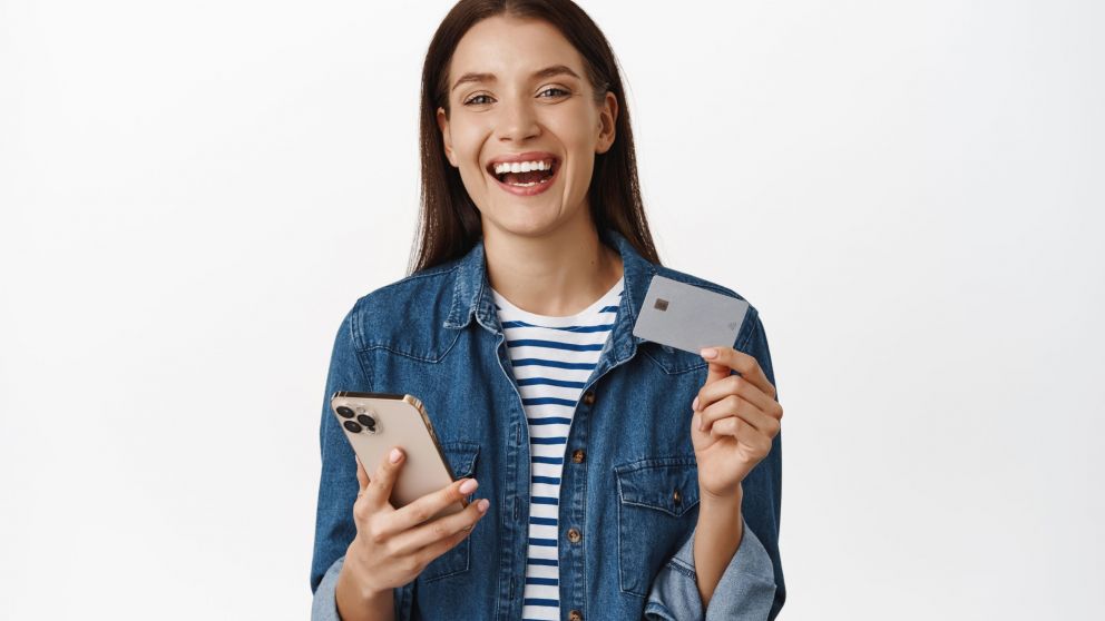 Happy woman holding smartphone, showing credit card, smiling satisfied and laughing, advertising online shopping sale, buying smth on mobile phone, white background.