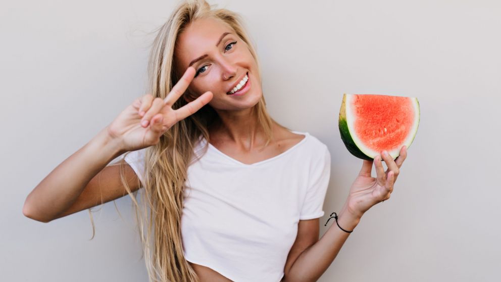 Amazing long-haired girl eating fruits and posing with peace sign. Indoor photo of carefree tanned woman with slice of watermelon.