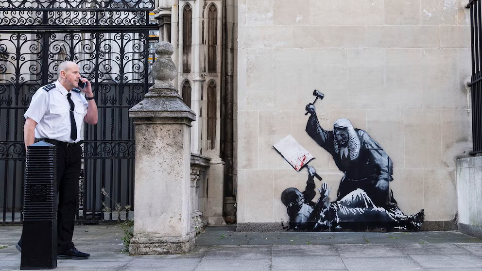 Banksy, aún anónimo, intervino el edificio de los tribunales reales de Londres con la imagen de un manifestante en el suelo, con un cartel ensangrentado, mientras un juez tenía en alza un martillo como arma